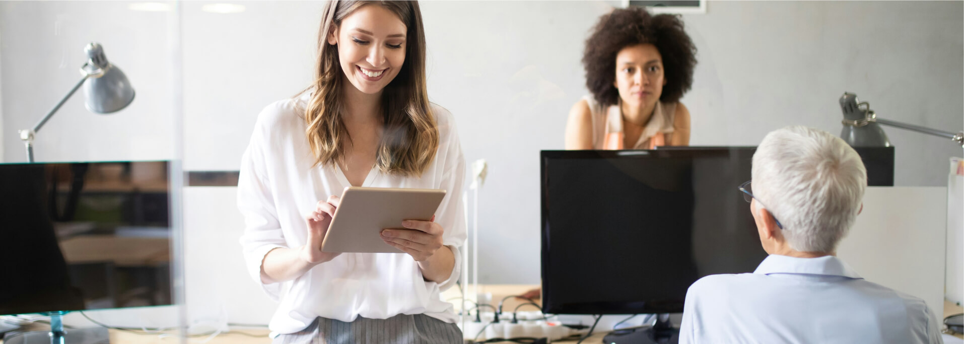 Drei Personen in einem modernen Büro, eine Frau lächelt und arbeitet mit einem Tablet, während die anderen im Gespräch sind.