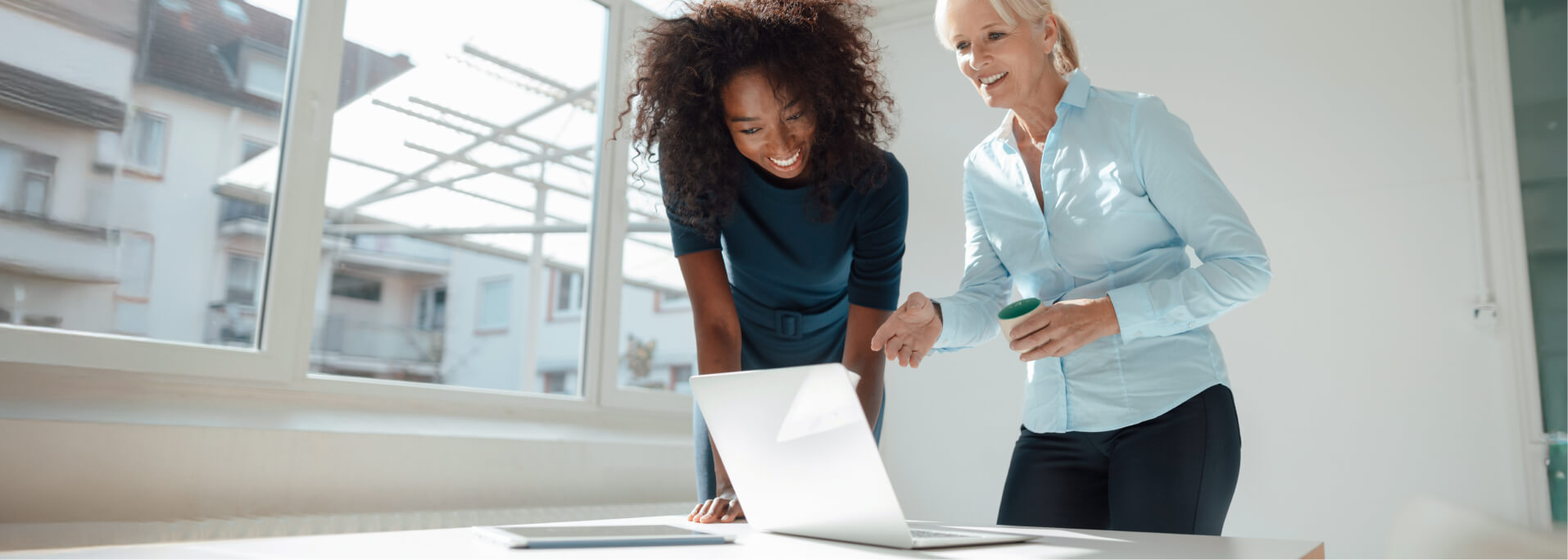 Zwei Frauen stehen lächelnd an einem Tisch und schauen gemeinsam auf einen Laptop.